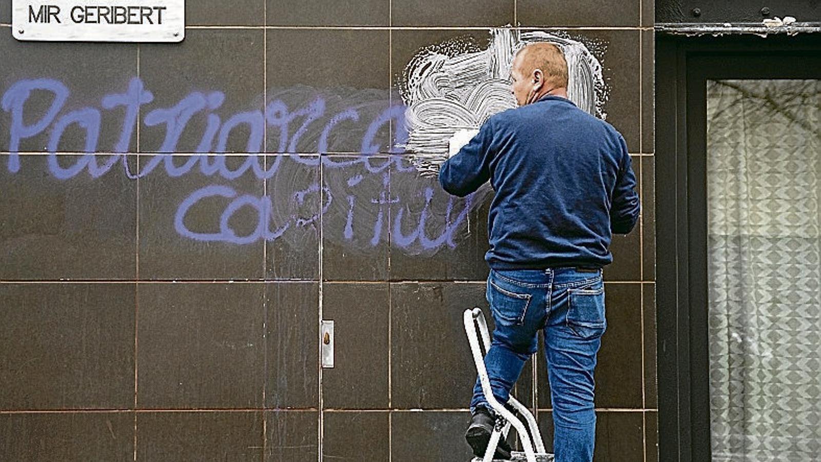 Un home netejant una pintada contra el patriarcat i el capital d’una paret d’un carrer de Barcelona.