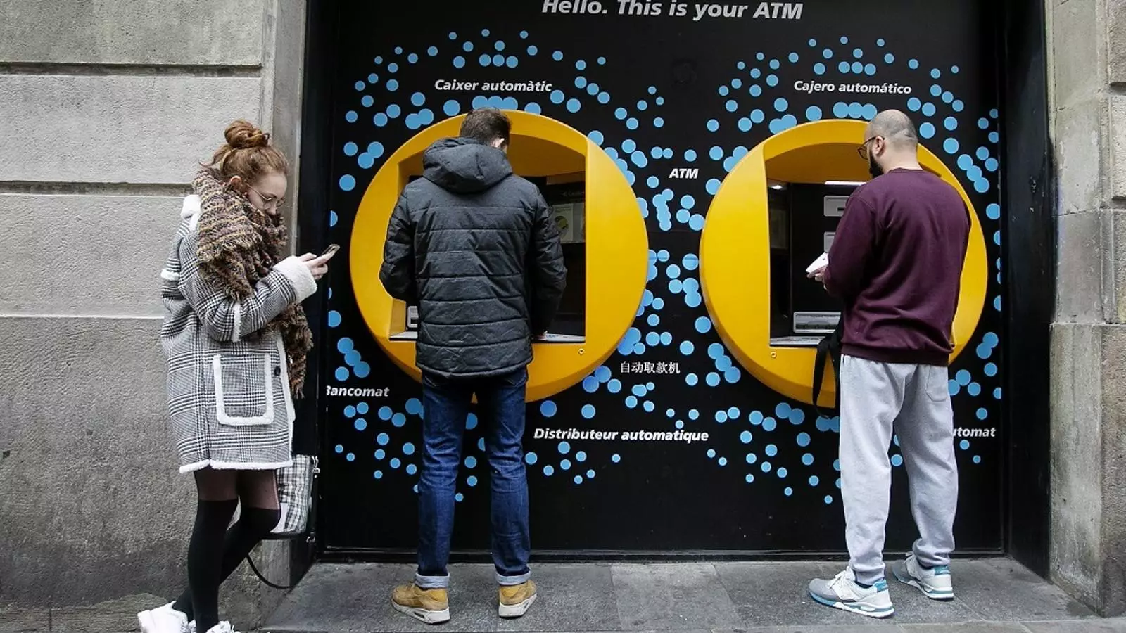 Dues persones fan servir un caixer automàtic a Barcelona, mentre una tercera s'espera, en una imatge d'arxiu.