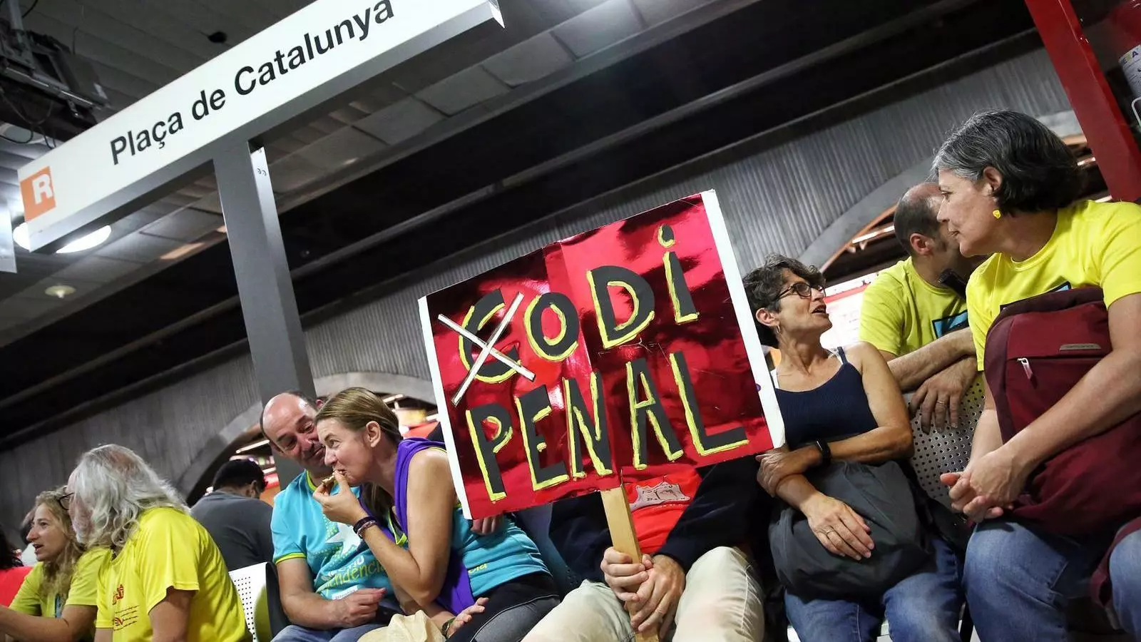 Gent esperant a l'andana de metro de Plaça de Catalunya. Una de les persones que s'hi espera porta una pancarta en la qual es pot llegir "Odi Penal" (fent el joc de paraules d'escriure 'Codi' i ratllant la 'c')