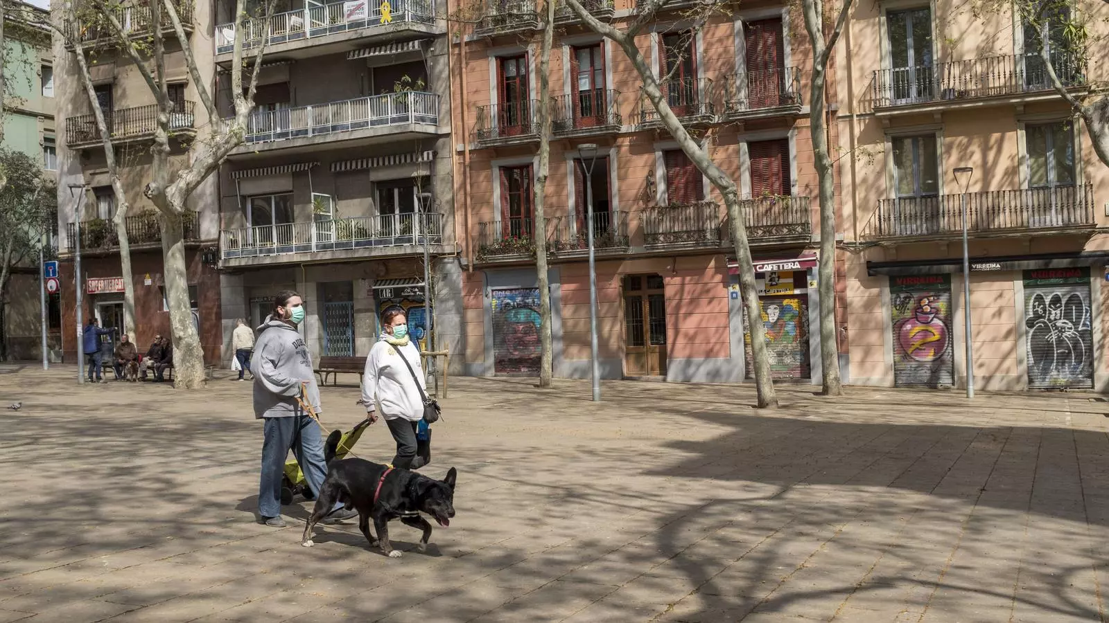 Dues persones passejant el gos a la plaça de la Virreina de Barcelona, amb els comerços tancats
