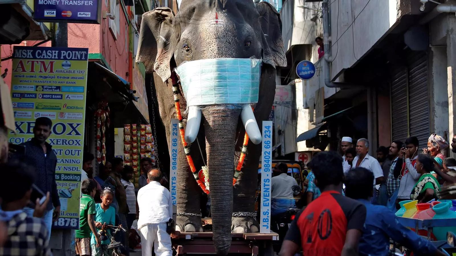 Un enorme elefant de cartró amb mascareta passant per un carrer ple de gom a gom a l'Índia