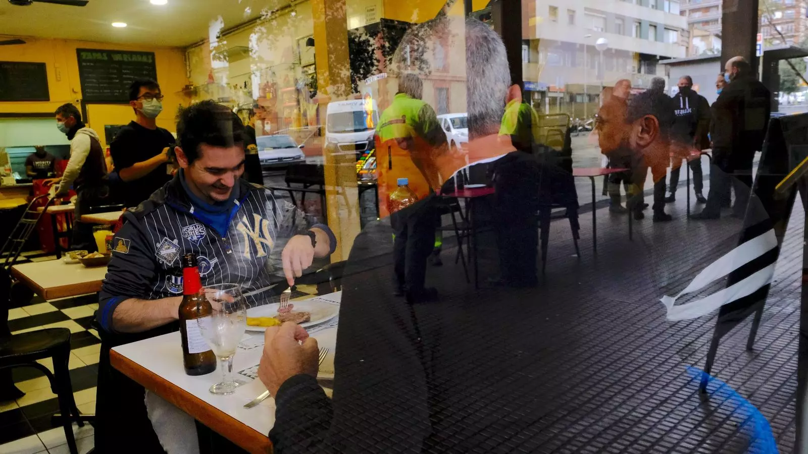 Tres comensals compartint taula a l'interior del bar amb el reflex de la terrassa exterior al finestral