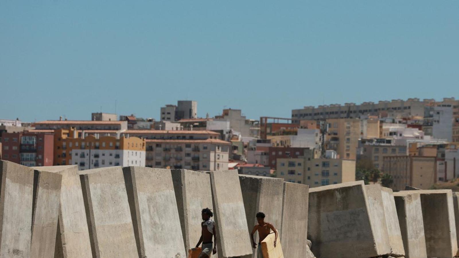 Two people with a mattress next to some jetties on a beach in Ceuta.
